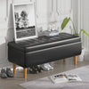 Black storage bench with wooden legs in a room setting with a plant and framed picture.