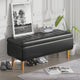 Black storage bench with wooden legs in a room setting with a plant and framed picture.